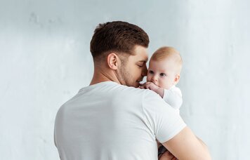 Ein männlicher Erwachsener hält ein Baby auf dem Arm und wendet ihm sein Gesicht lächelnd zu. | © Symbolbild: AdobeStock #360042512 | LIGHTFIELD STUDIOS