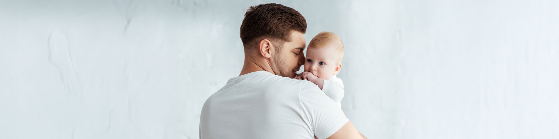 Ein männlicher Erwachsener hält ein Baby auf dem Arm und wendet ihm sein Gesicht lächelnd zu. | © Symbolbild: AdobeStock #360042512 | LIGHTFIELD STUDIOS